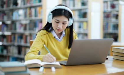 a person with black hair wearing a yellow top and pale blue headphones sits at a desk with a laptop and open books